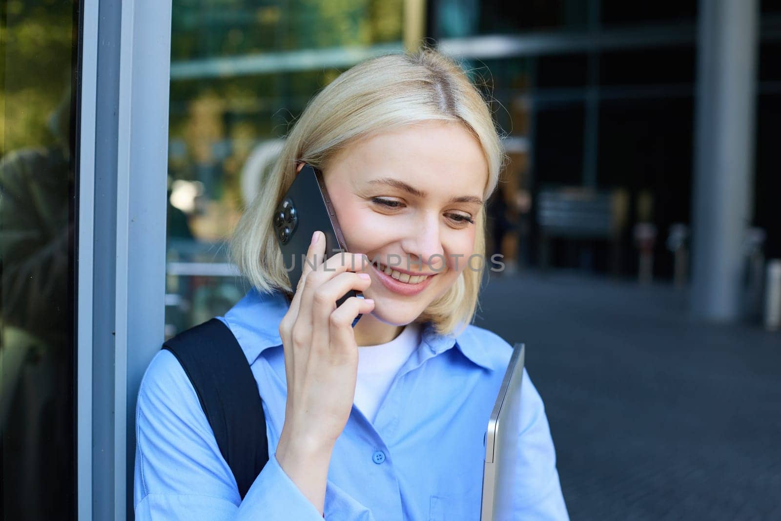 Happy female model, blond woman talking on mobile phone, chatting with someone, standing near office building with laptop and smartphone, smiling.