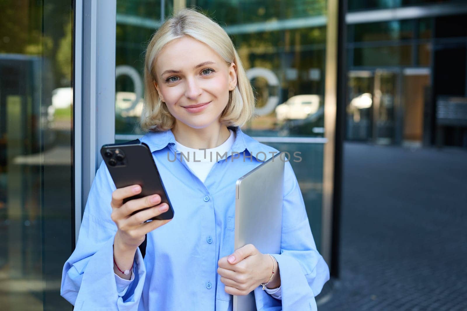 Portrait of young female student standing outside university campus, posing near building, holding laptop and smartphone, wearing blue collar shirt by Benzoix