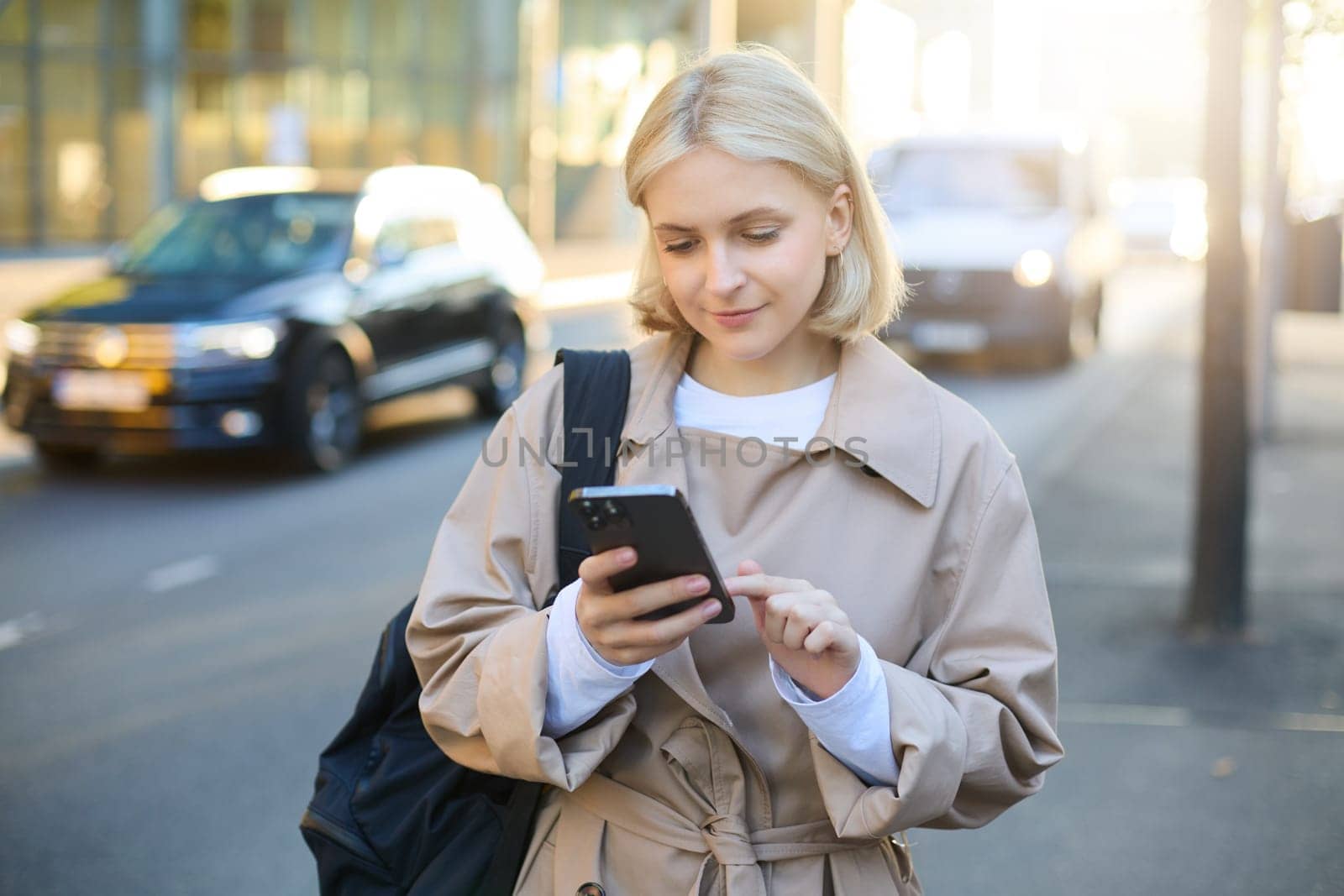 Close up portrait of young modern woman on street, using mobile phone, looking at smartphone screen, reading notification, waiting for taxi near road by Benzoix