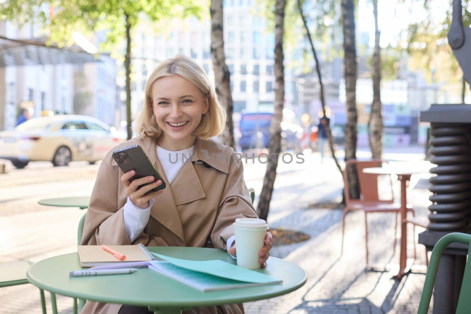 Portrait of blond smiling woman with smartphone, holding cup of coffee, drinking chai and enjoying sunny day outdoors in city centre by Benzoix