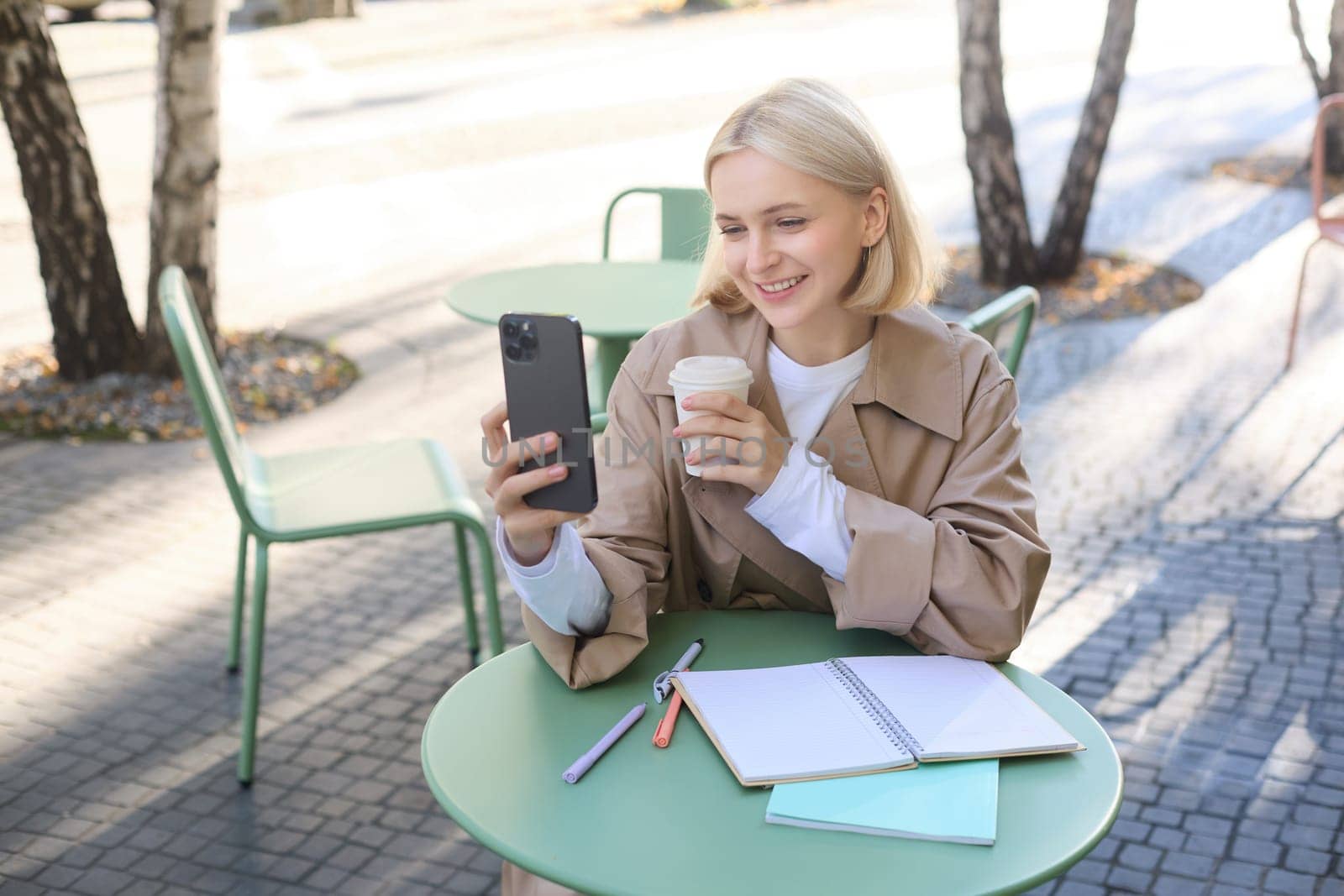 Lifestyle shot of stylish blond girl with smartphone, drinking coffee, posing with her chai for social media, creating content on phone, taking selfie.