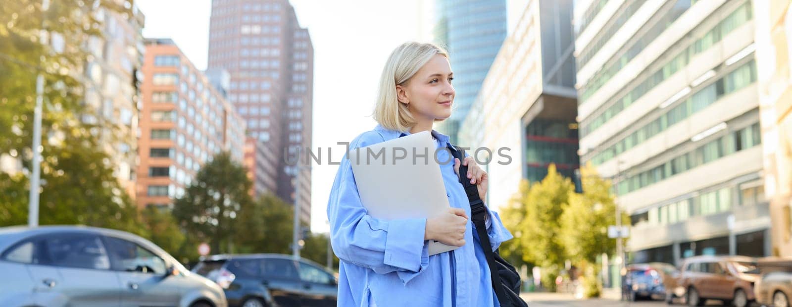 Street stylish portrait of young happy woman, blond girl with backpack and laptop, standing outdoor s near road on sunny bright day by Benzoix