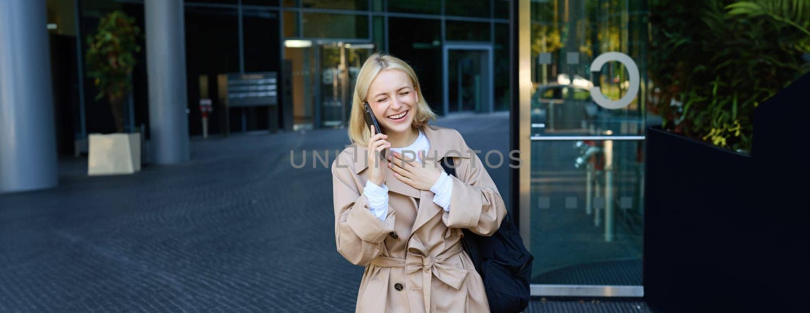 Carefree blond female model, talking to friend on mobile phone, waiting for someone near building on street, chatting on smartphone and laughing.