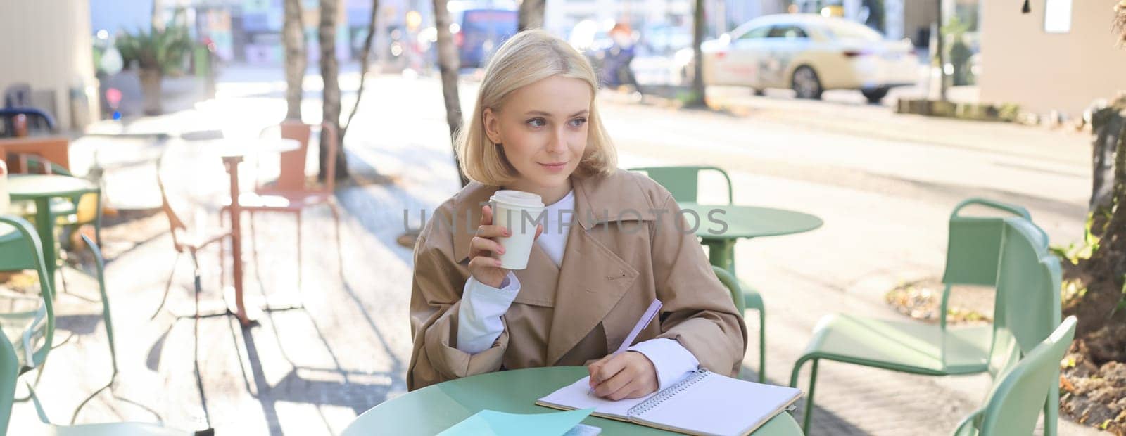 Portrait of beautiful blond girl, college student writing in notebook, doing homework in outdoor cafe, drinking coffee, smiling and looking at the street by Benzoix