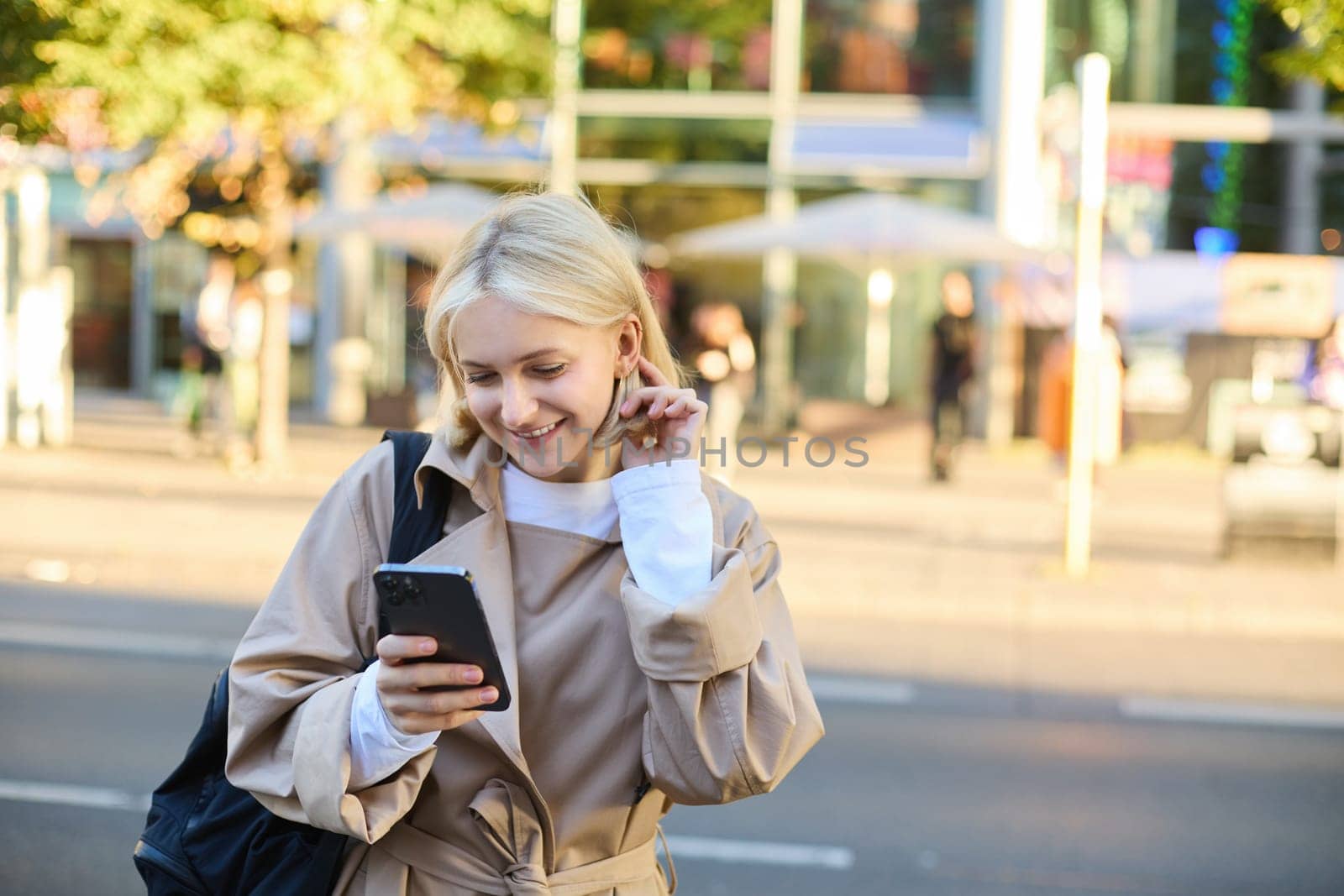 Modern young woman on street, reading message on mobile phone, using smartphone, standing near busy road and smiling by Benzoix