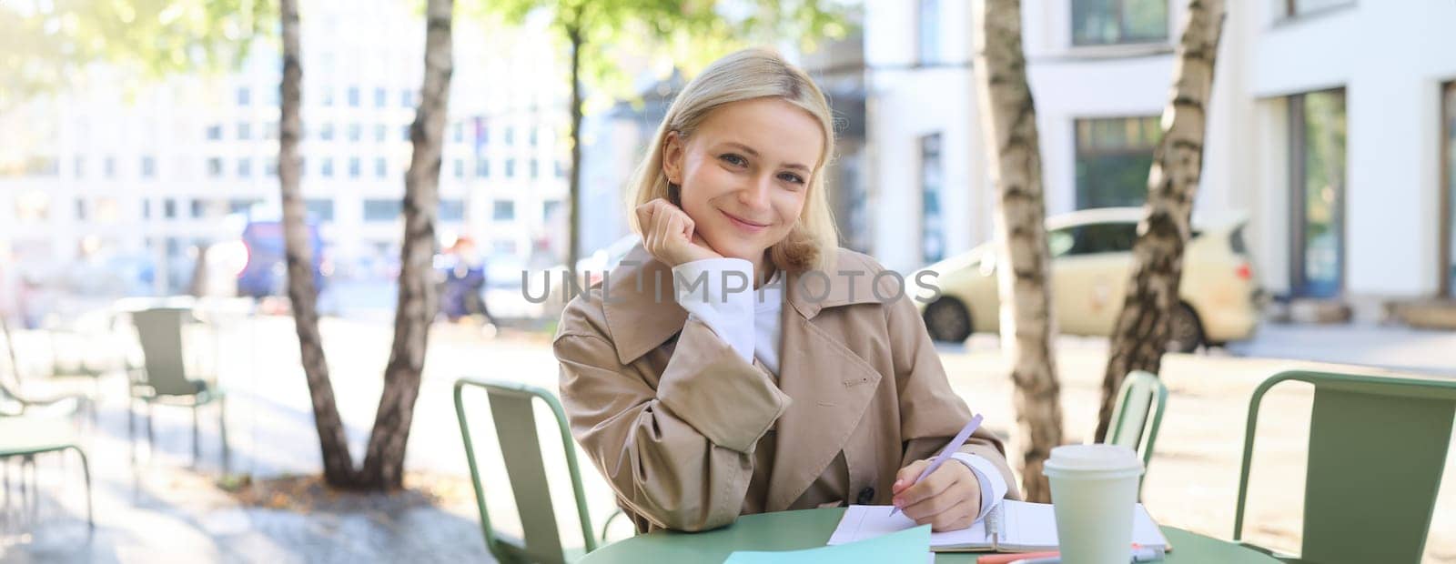 Portrait of young modern woman in stylish trench coat, sitting in outdoor cafe on sunny day, writing in her notebook, working or doing homework, drinking coffee.