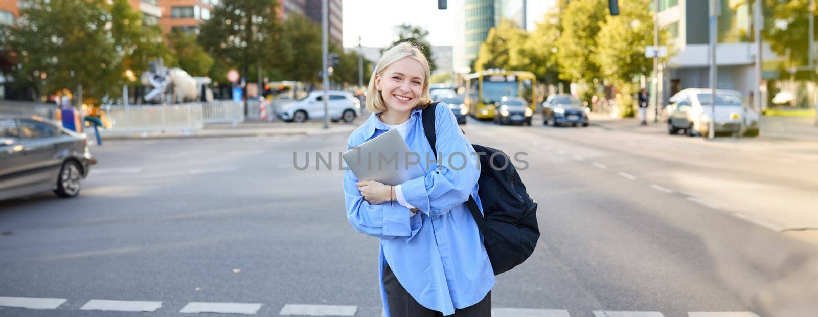 Image of young college student, happy female model on street, posing with backpack and laptop outdoors, with roads and cars behind her back, smiling at camera by Benzoix