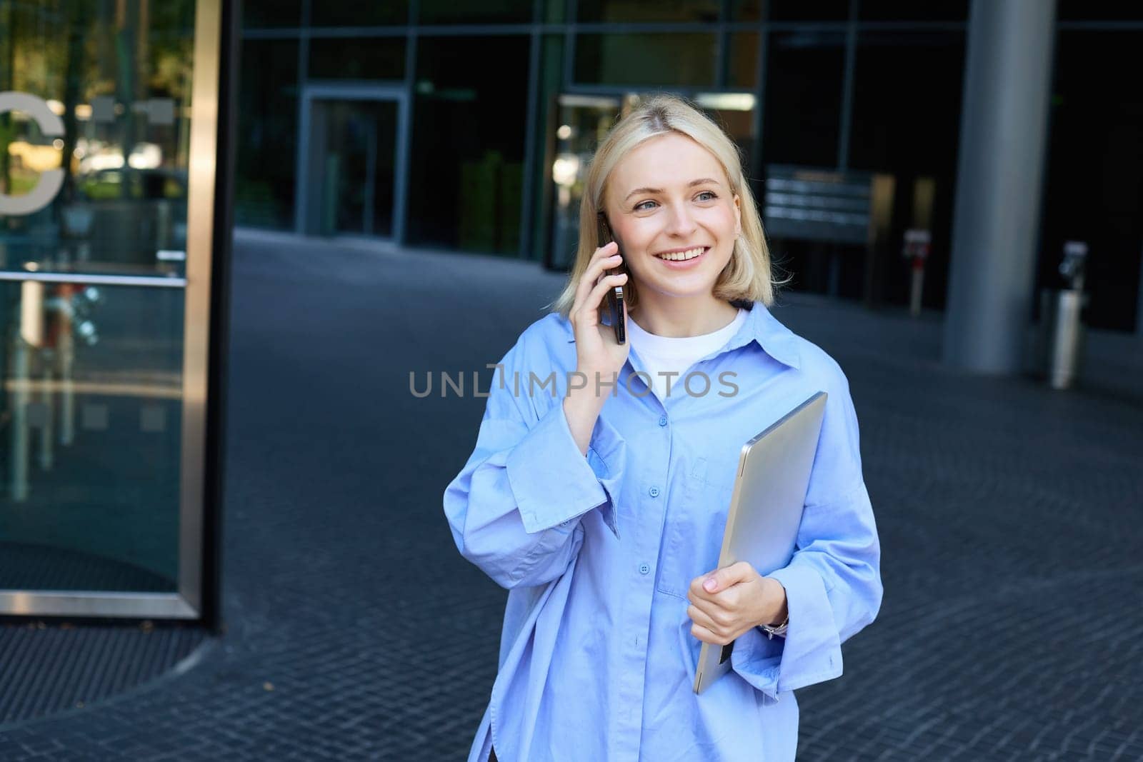 Image of young smiling woman, working in company, standing near office with laptop, answers phone call, chatting with someone by Benzoix