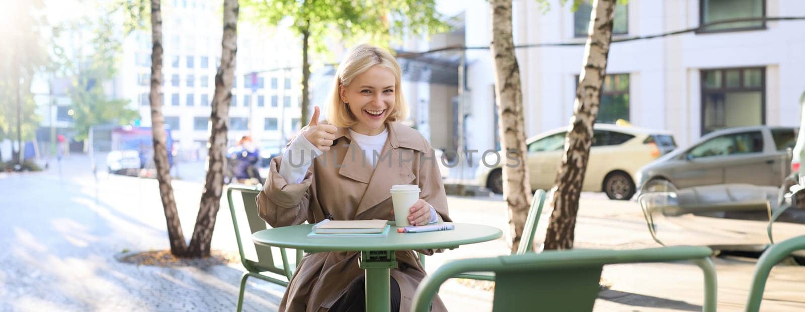 Smiling happy woman, sitting in cafe and showing thumbs up, recommending coffee place, gives her positive feedback.
