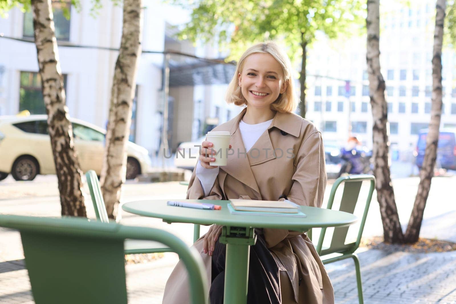 Portrait of young blonde woman sitting in cafe, doing homework and drinking coffee, writiing in journal, using notebook on fresh air by Benzoix
