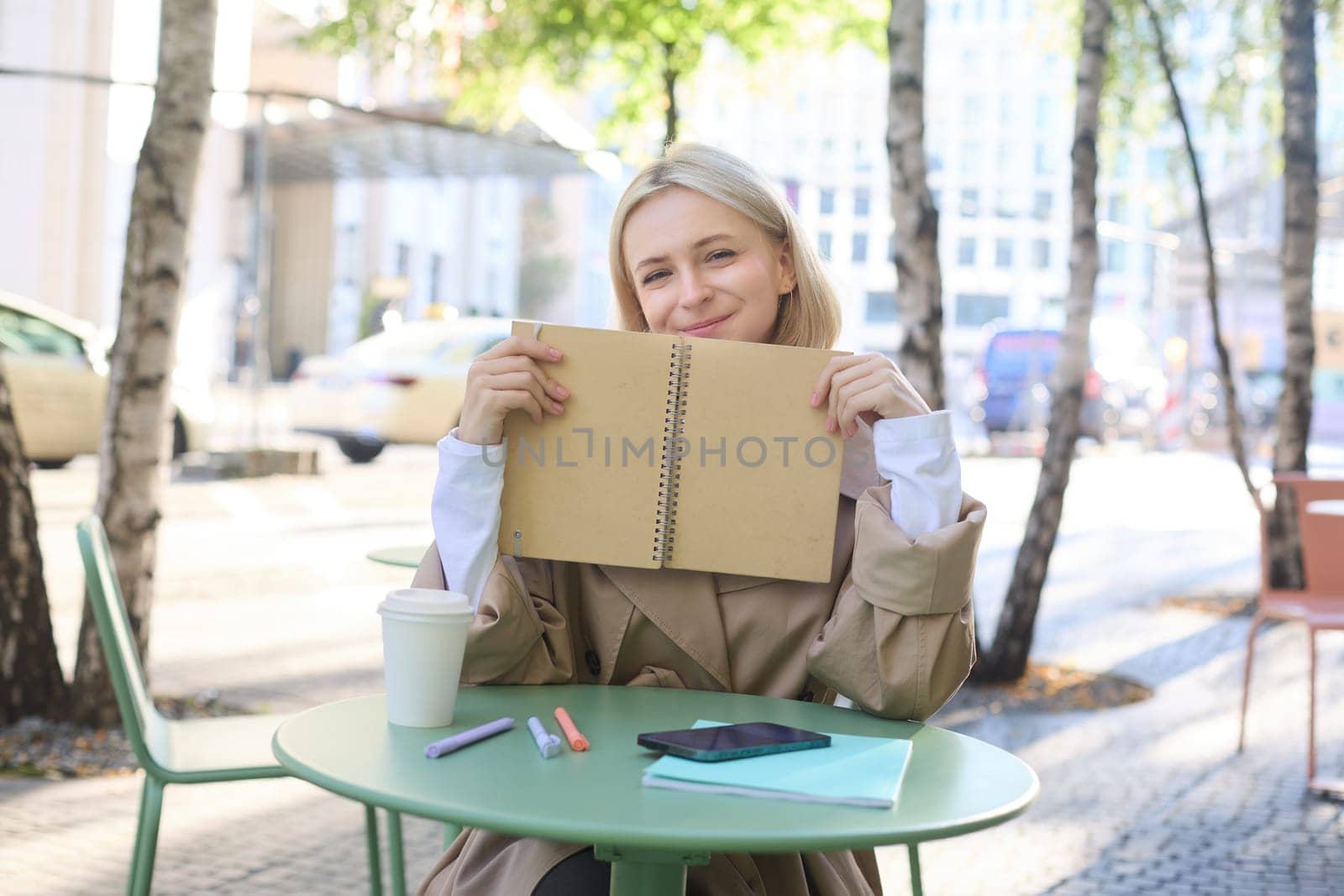 Portrait of young woman sitting in outdoor cafe, hiding behind journal, holding notebook in hands and smiling by Benzoix
