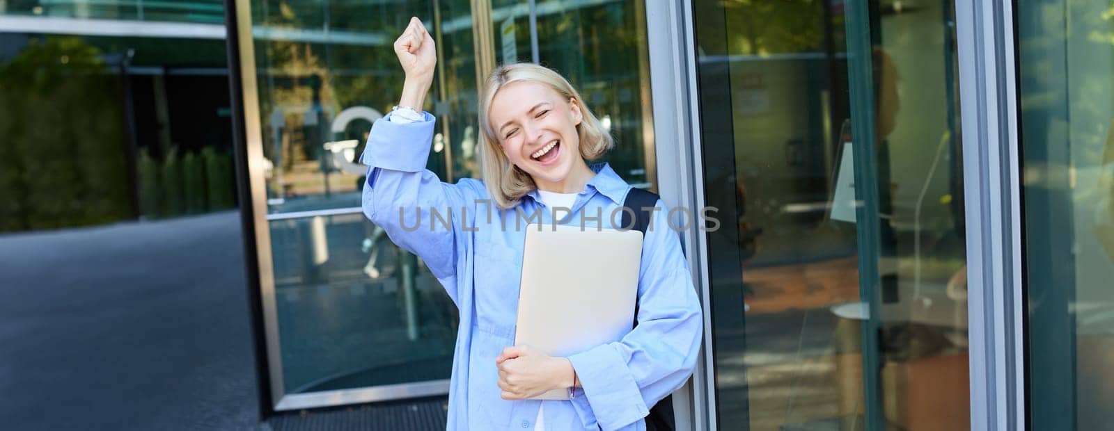 Carefree blond woman laughing, smiling and celebrating, posing with laptop near office, campus building, raising hand up in triumph, feeling excited by Benzoix