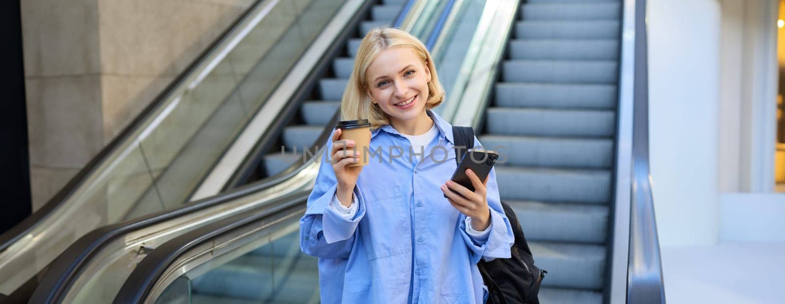 Image of beautiful female model, student standing near escalator, raising cup of takeaway coffee, smiling and looking happy, has mobile phone in hand and backpack on shoulder.