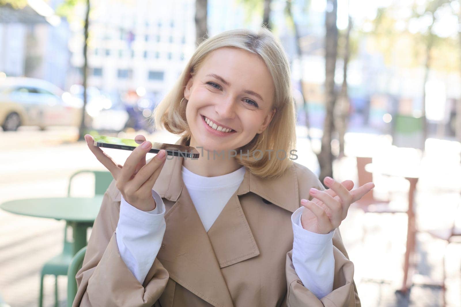 Close up portrait of smiling blond woman, girl with mobile phone outdoors, sitting in cafe, recording voice message, talking into smartphone with microphone near mouth by Benzoix