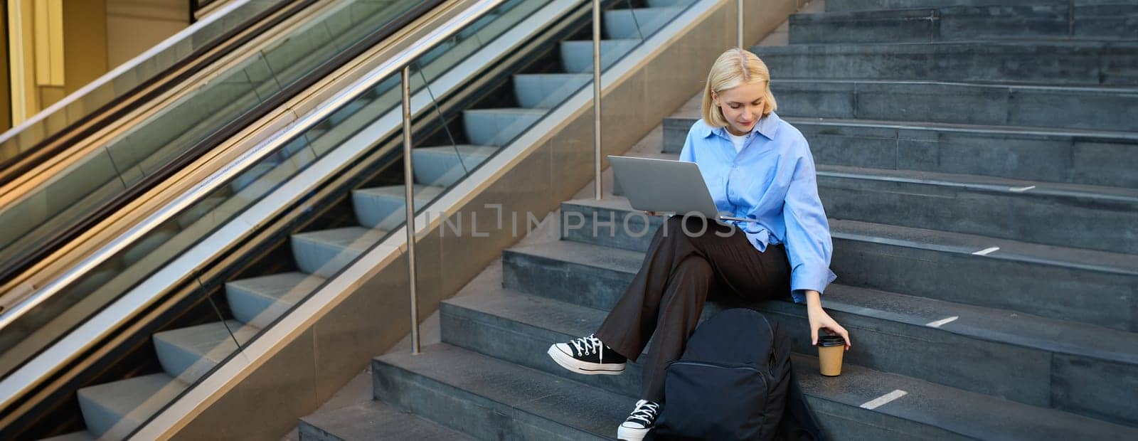Modern young woman, student working on laptop, sitting outside campus in stairs with backpack and takeaway coffee, drinking her beverage and studying by Benzoix