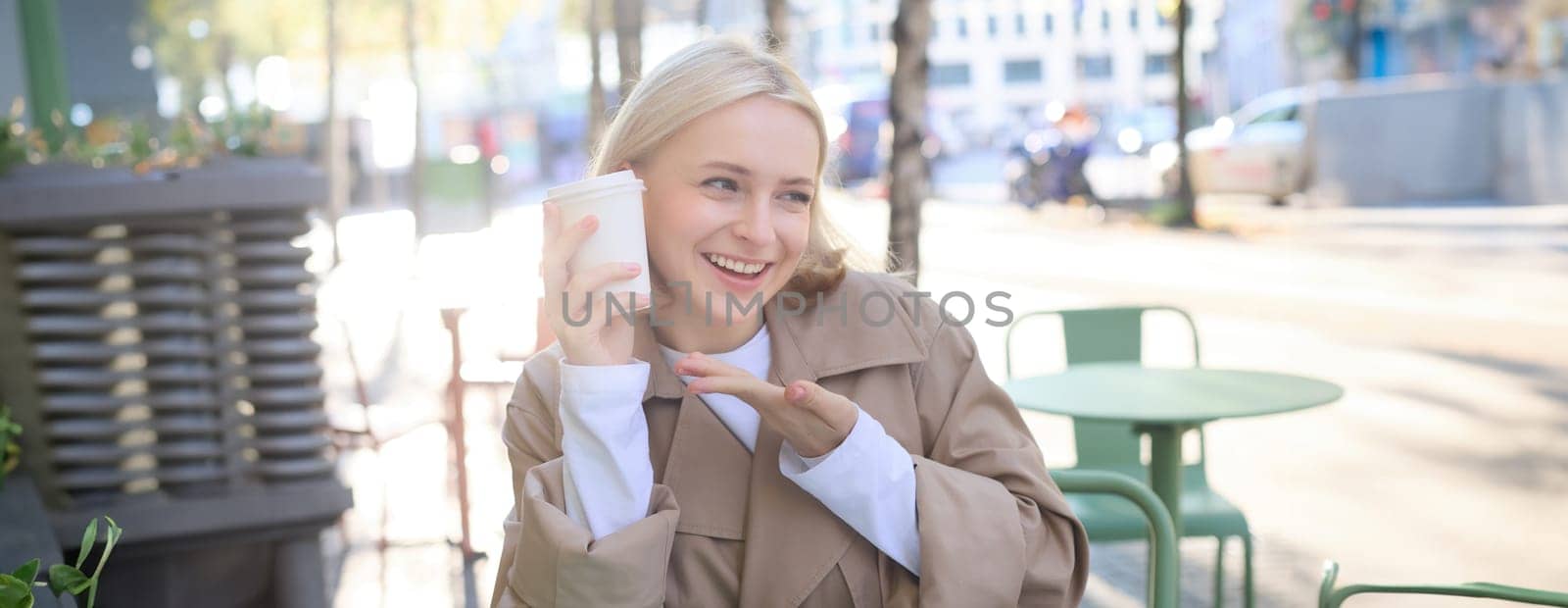 Portrait of smiling blond woman, pointing finger at her cup of coffee, orders takeaway, sits in street cafe, shows her favourite drink by Benzoix