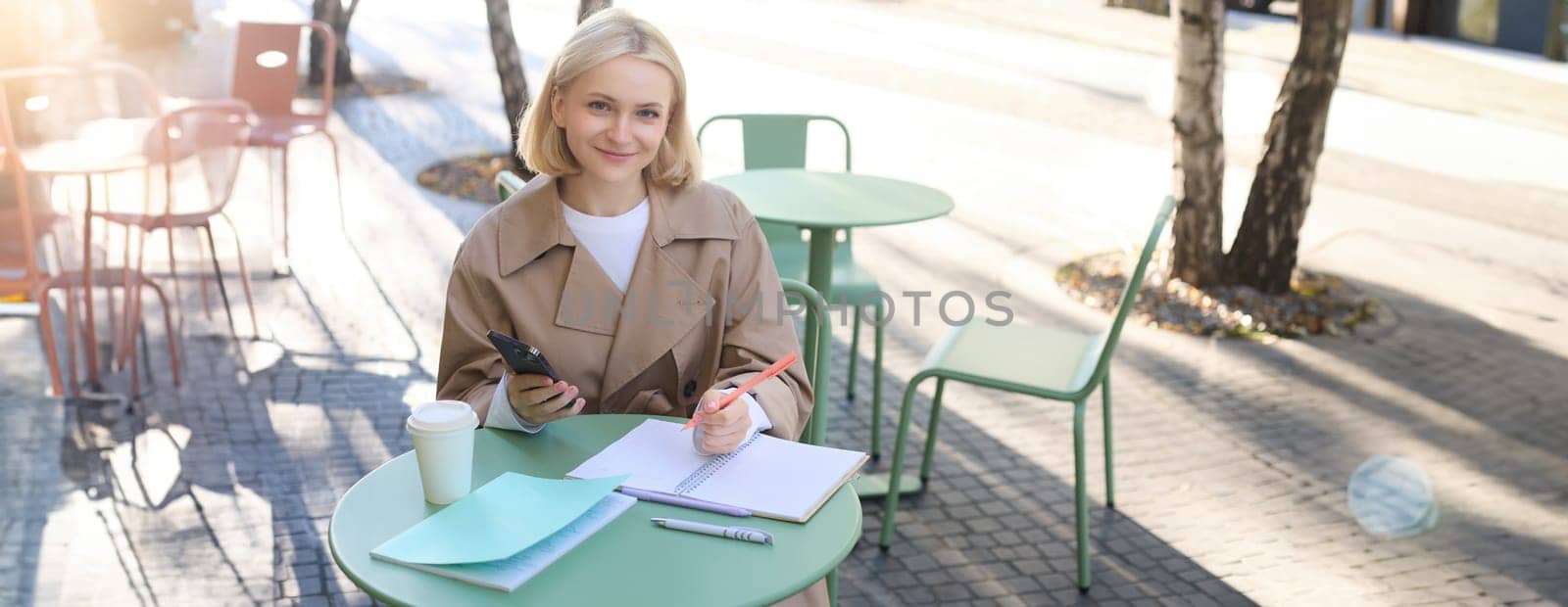 Happy beautiful young woman enjoying the sun outdoors, sitting in city centre cafe, drinking coffee, holding smartphone and laughing, smiling with eyes closed.