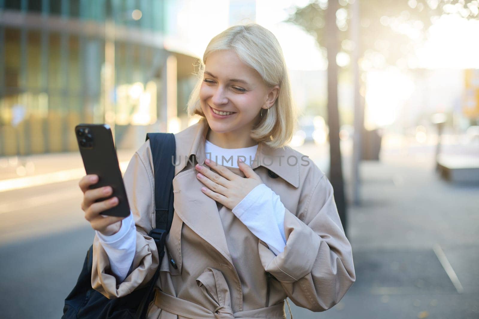 Image of stylish female model with smartphone, looking at mobile phone, video chatting, talking to someone online, pointing at herself and smiling, walking on street.