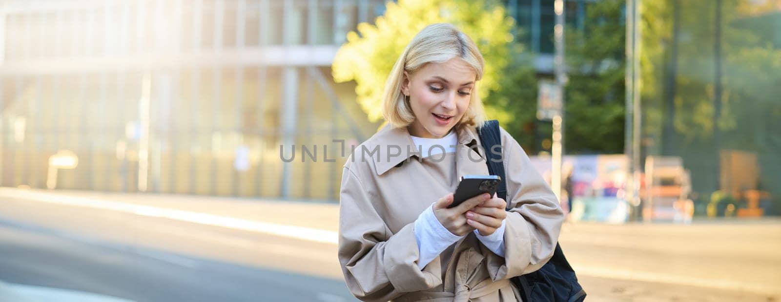 Portrait of surprised girl on street, woman looking at her phone with raised eyebrows, reading a message on smartphone, waiting for someone near the road, holding backpack by Benzoix