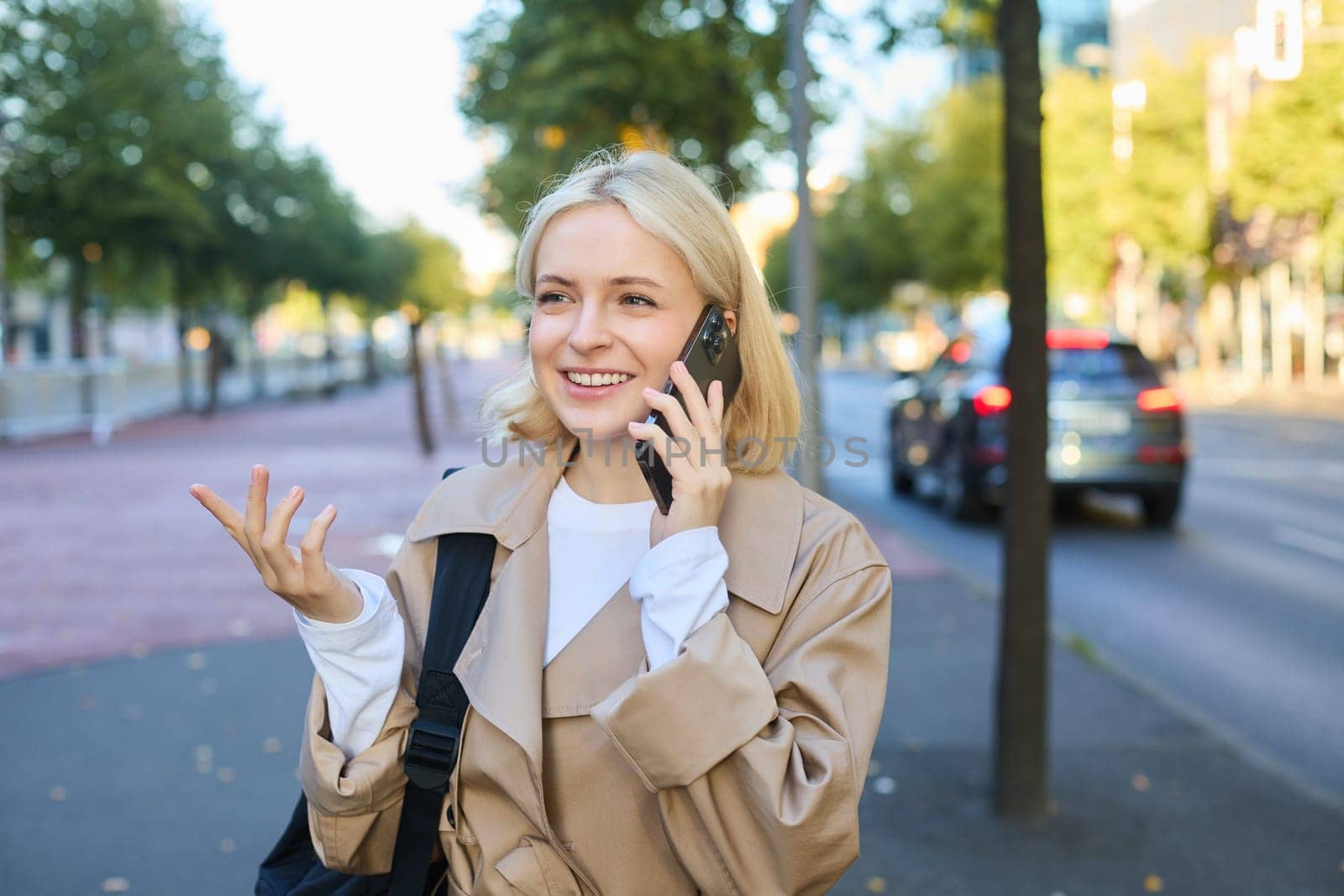 Close up portrait of beautiful young woman, blonde girl walking on street with mobile phone, chatting with friend, has happy face expression while talking over cellphone on her way.