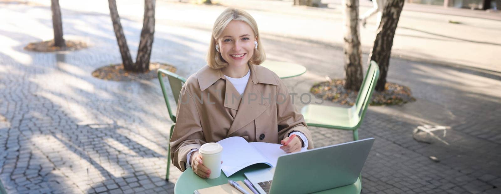 Portrait of beautiful blond woman, wearing wireless headphones, using laptop, studying in outdoor coffee shop, making notes, working on project.