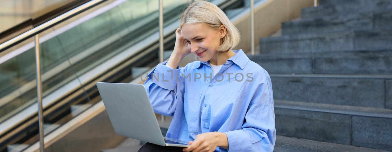 Young woman, student in blue shirt, working on laptop, sitting outdoors on street stairs, working on project online, connects to public wifi, elearning.