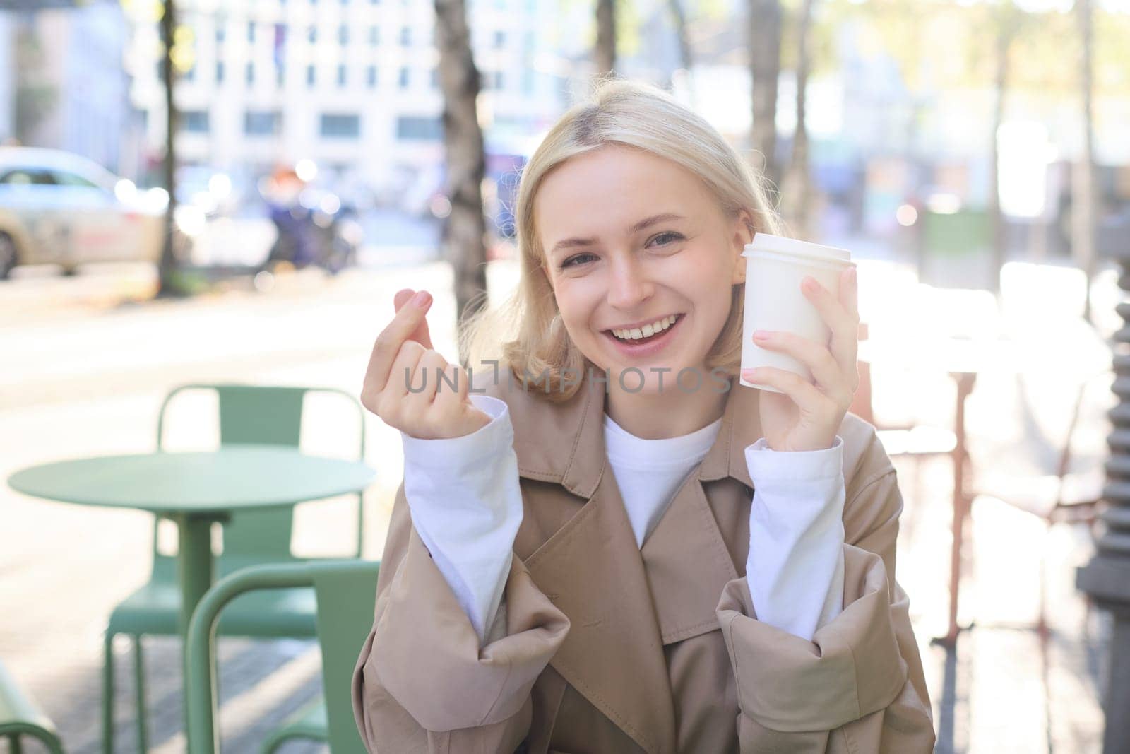 People and leisure concept. Happy blonde girl, sitting with cup of coffee in street cafe, showing finger heart sign, enjoys spending time outdoors by Benzoix