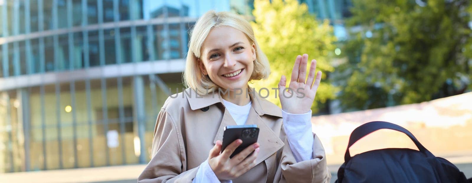 Portrait of young blonde college girl, student sitting on street bench, holding smartphone, seeing a friend and waving hand to say hello, smiling friendly at camera.