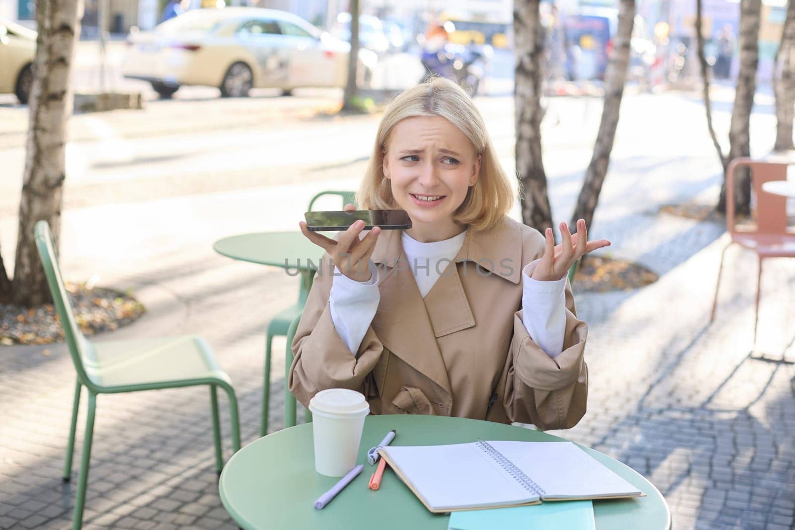 Portrait of young urban female model, sitting in cafe outdoors, recording voice message with upset, confused face, shrugging, holding mobile phone near mouth by Benzoix