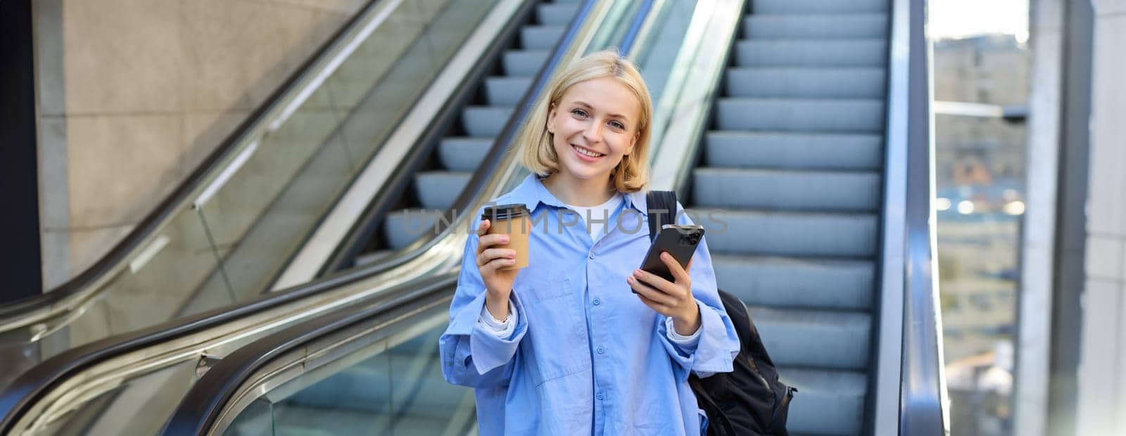 Image of stylish young woman in blue shirt, holding takeaway cup of coffee and mobile phone, standing near escalator and waiting for you, smiling at camera by Benzoix