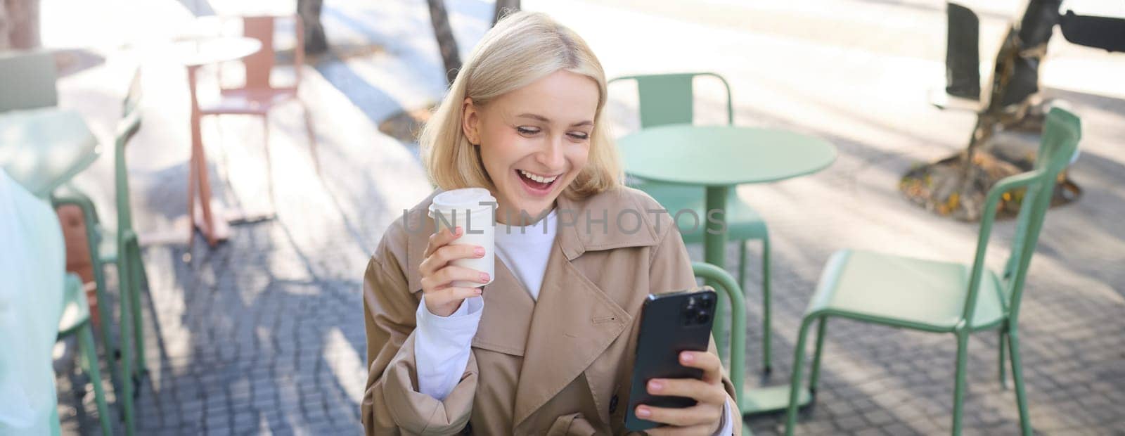 Lifestyle portrait of smiling young woman, sitting outdoors with coffee drink, looking at smartphone with happy, excited face expression. Technology and people concept