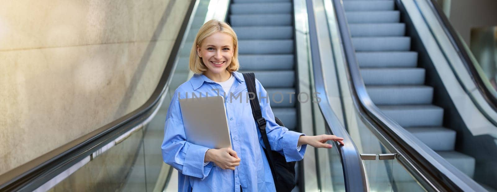 Street style portrait of woman on escalator, holding laptop and backpack on her shoulder, smiling at camera, walking in city.