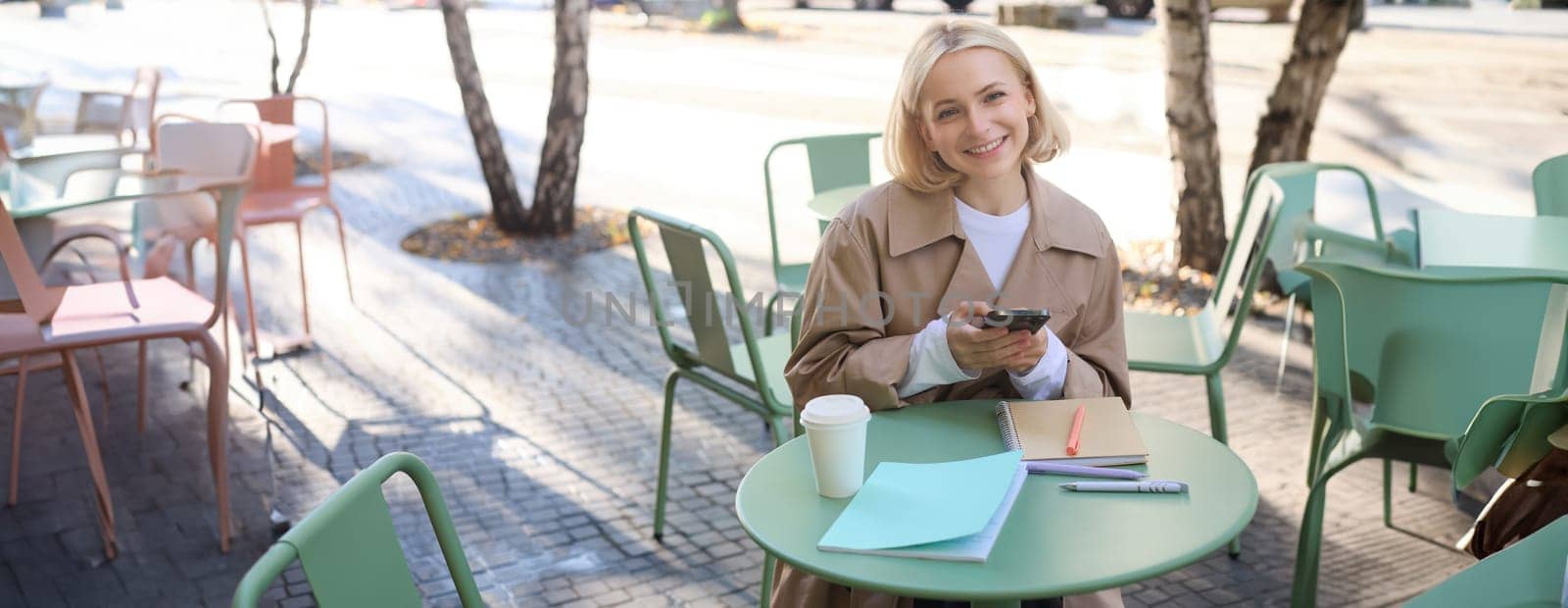 Portrait of cute smiling woman with blond short hair, sitting with documents and mobile phone, looking happy at camera by Benzoix