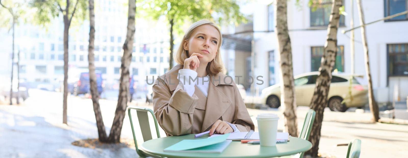 Portrait of young woman thinking while doing homework, sitting in outdoor cafe alone, drinking coffee and writing in notebook, looking thoughtful, pondering smth by Benzoix