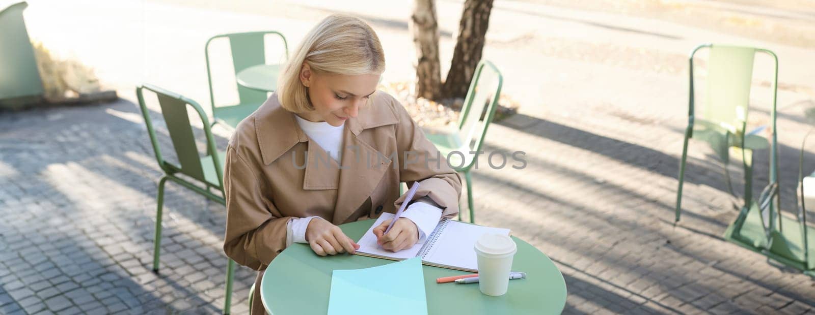 Portrait of woman working on project, writing in notebook, sitting outdoors in cafe, drinking coffee and doing homework. Student lifestyle and people concept