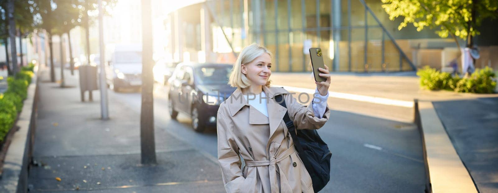 Image of stylish smiling woman, blogger taking selfie on street, smiling and posing for social media photo on smartphone, using mobile phone camera.