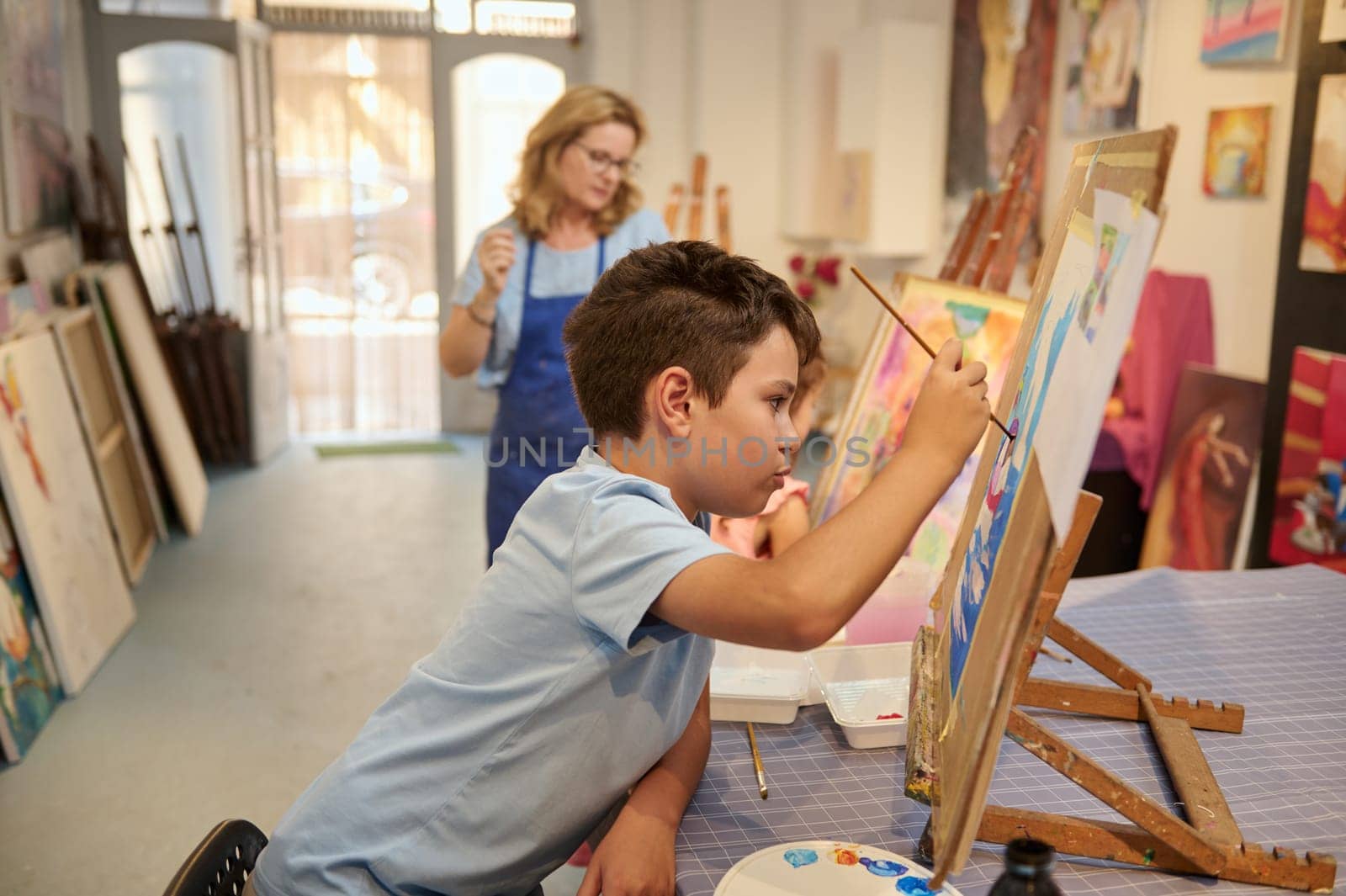 Authentic side portrait of a Caucasian handsome preteen school boy artist holding a paintbrush, focused on painting on easel in a creative art studio. Fine art. Drawing. Kids education concept