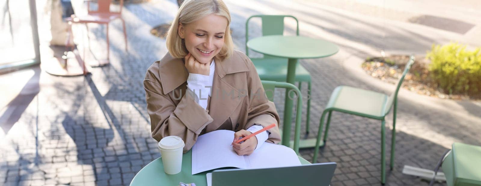 Image of smiling young female student, working on project, sitting outside on sunny day in cafe, looking at laptop and making notes, studying, doing homework outdoors by Benzoix