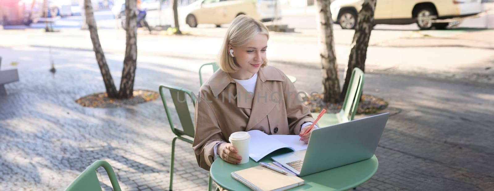 Lifestyle and people concept. Woman in outdoor cafe, sitting with laptop on street, connects to online course or lecture, doing homework, making notes, wearing wireless headphones, drinking coffee.