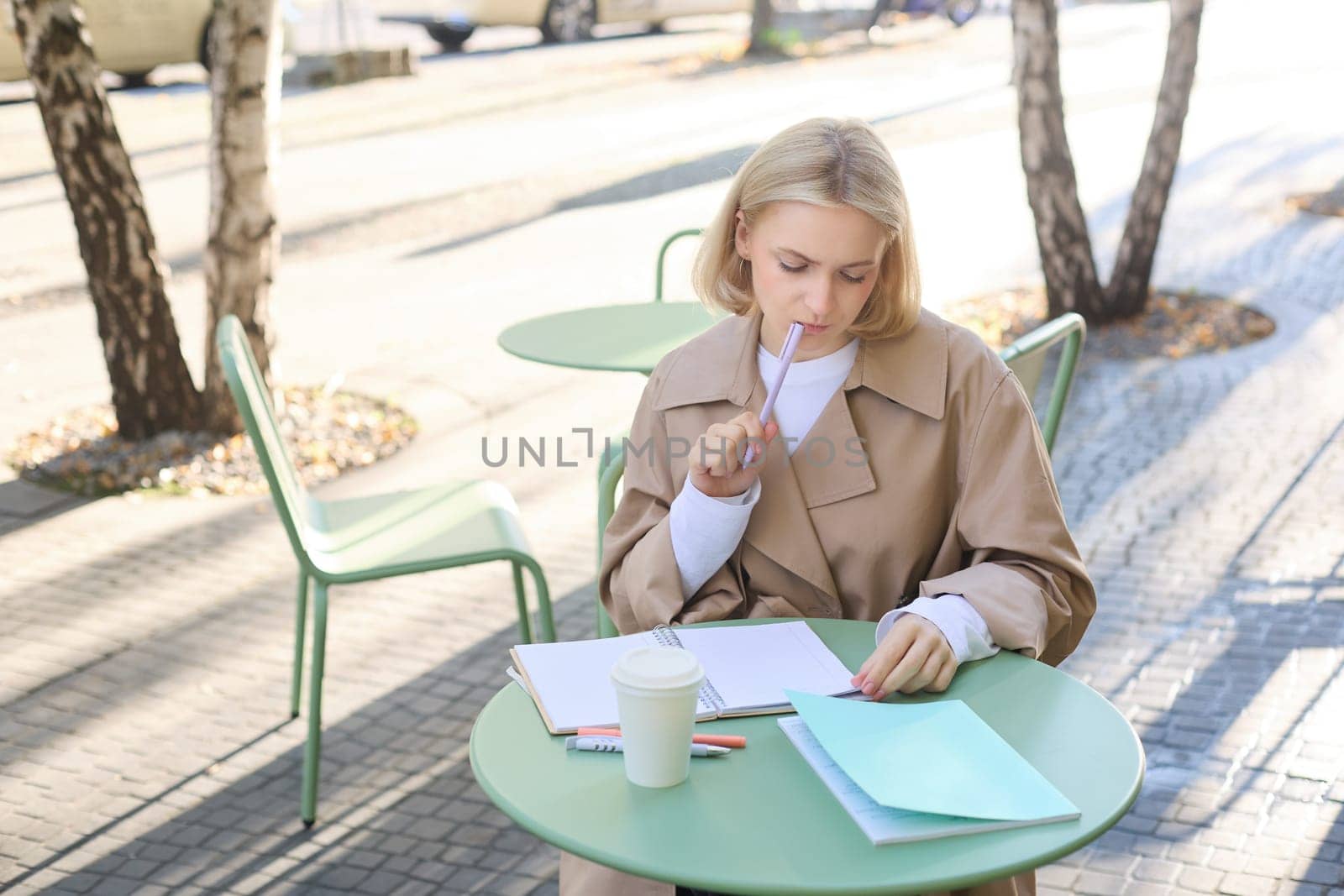 Portrait of young woman thinking while doing homework, sitting in outdoor cafe alone, drinking coffee and writing in notebook, looking thoughtful, pondering smth.