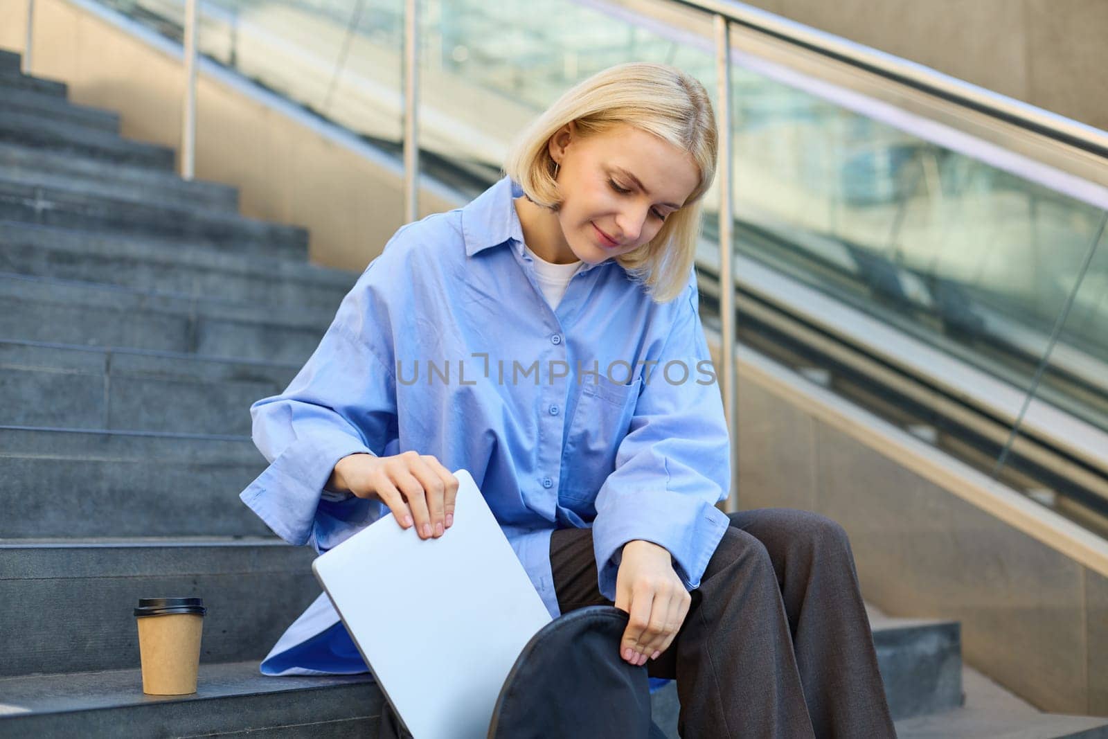 Smiling young female model, packing her laptop in backpack, sitting on city stairs outdoors, drinking cup of coffee before lecture in university by Benzoix