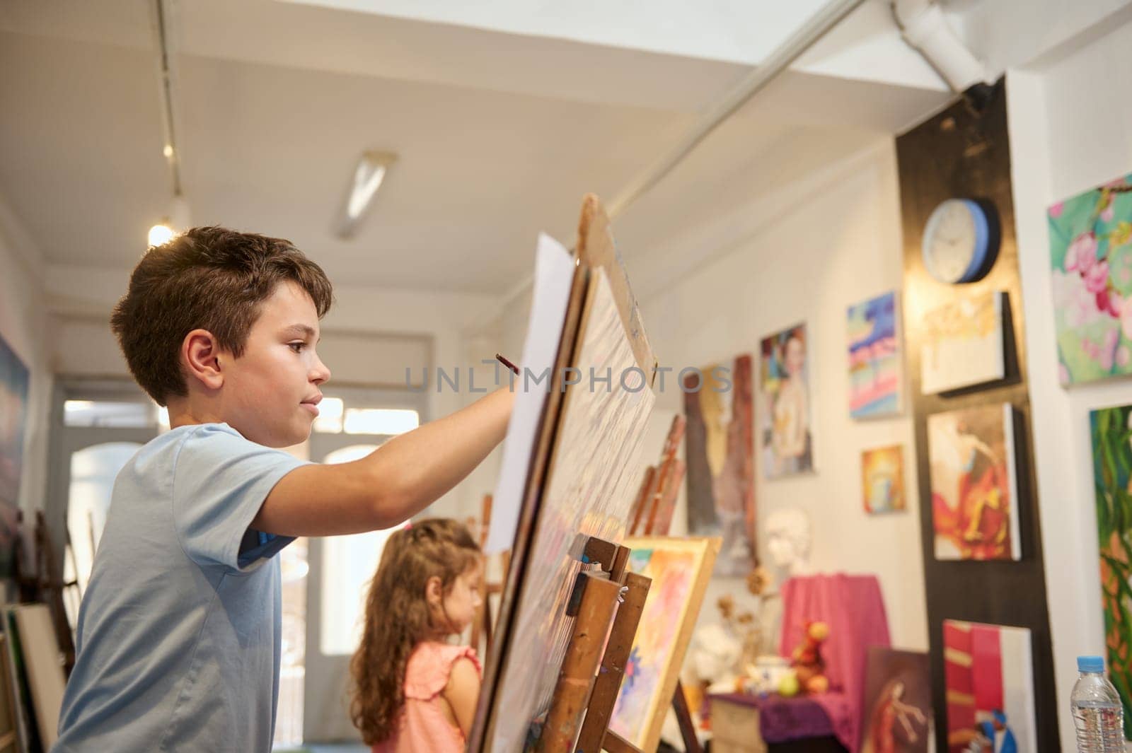 Lifestyle portrait of adorable talented kids learning drawing on canvas in a modern creative fine art studio. Children at art class in workshop with pictures exhibited on a wall in art gallery