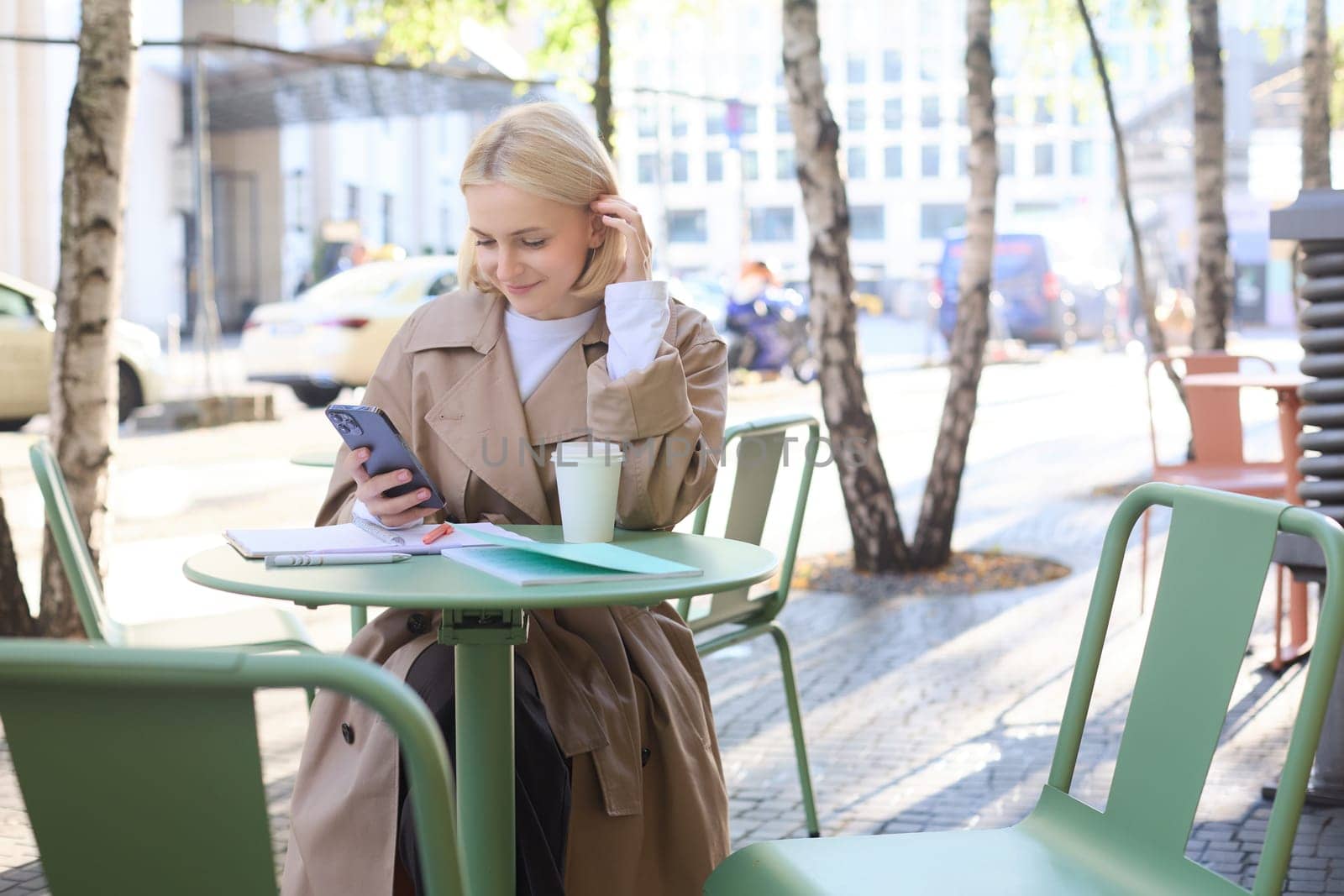 Street style portrait of young smiling woman sitting on street outdoors, holding mobile phone, using smartphone, drinking coffee in local cafe.