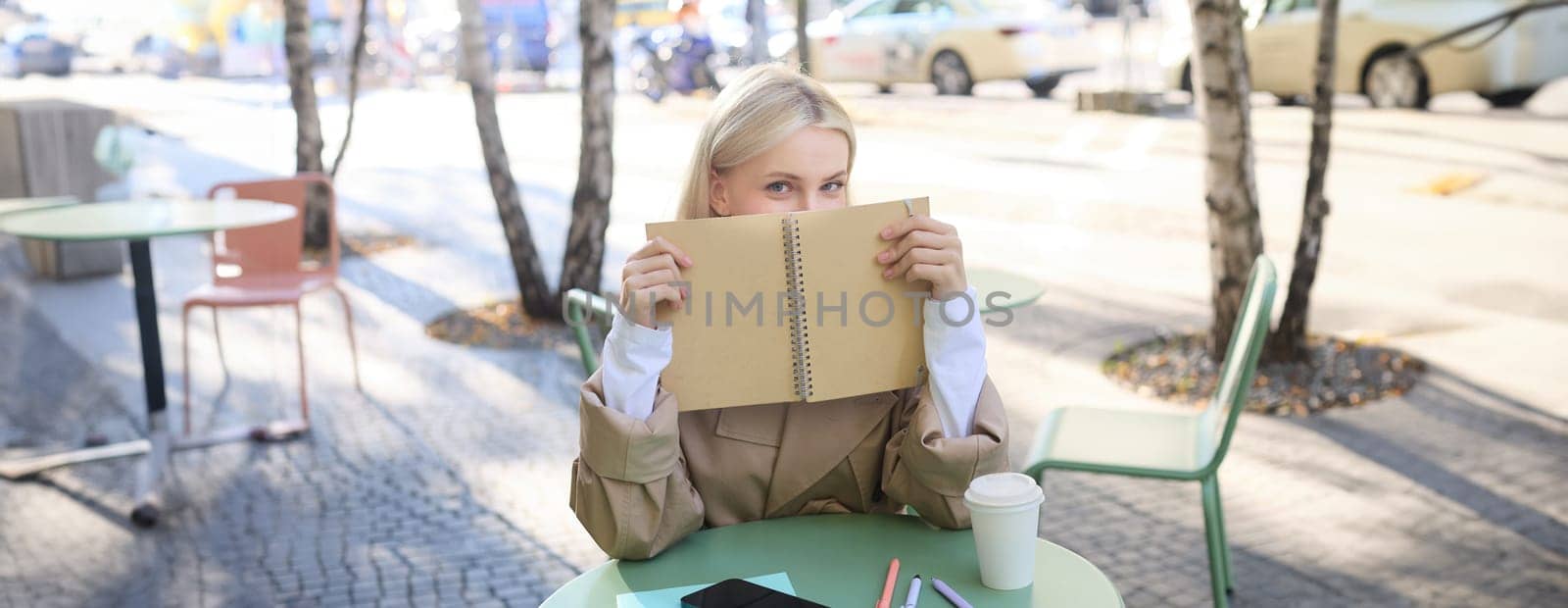 Portrait of young woman sitting in outdoor cafe, hiding behind journal, holding notebook in hands and smiling by Benzoix