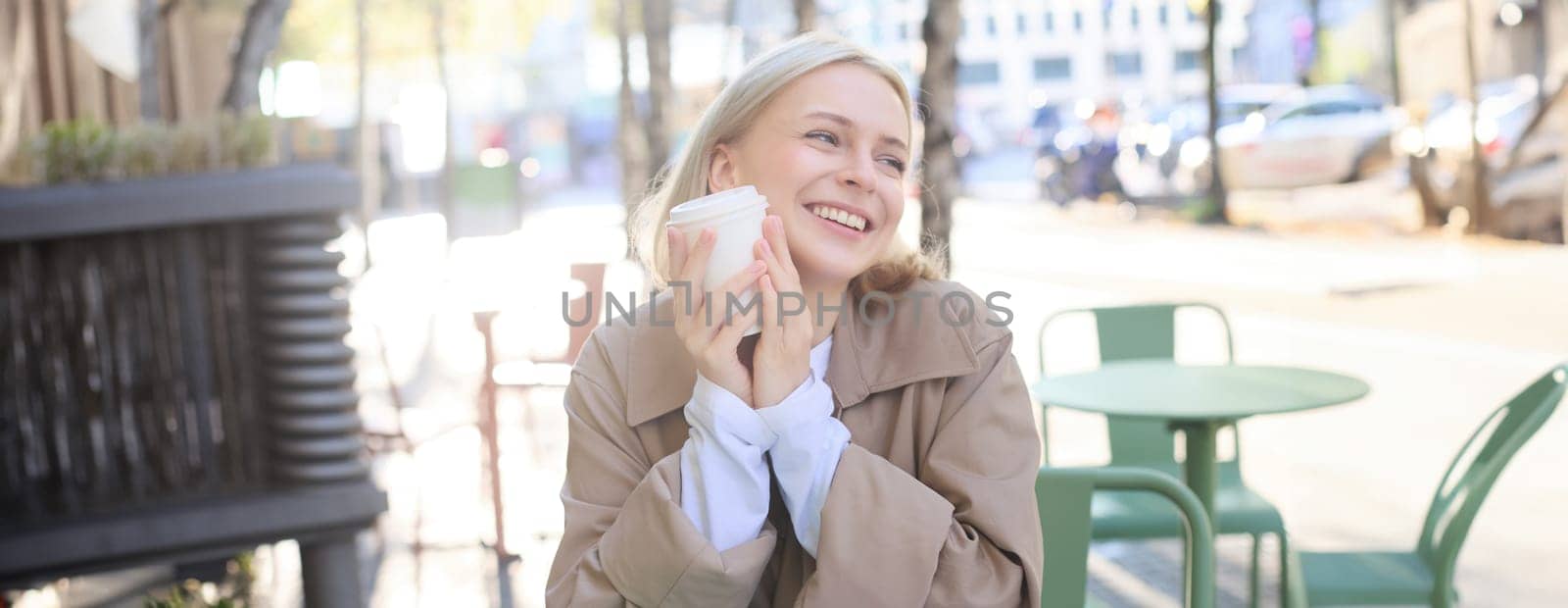 Portrait of cheerful young blond woman, enjoying spending time in outdoor cafe, drinking coffee and laughing, having casual meet up in street restaurant. Lifestyle concept