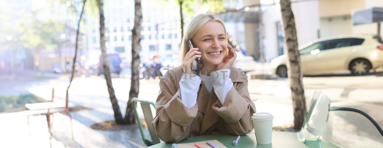 Close up portrait of smiling blond woman, sitting at table outside cafe, talking on mobile phone, having happy, lively chat by Benzoix