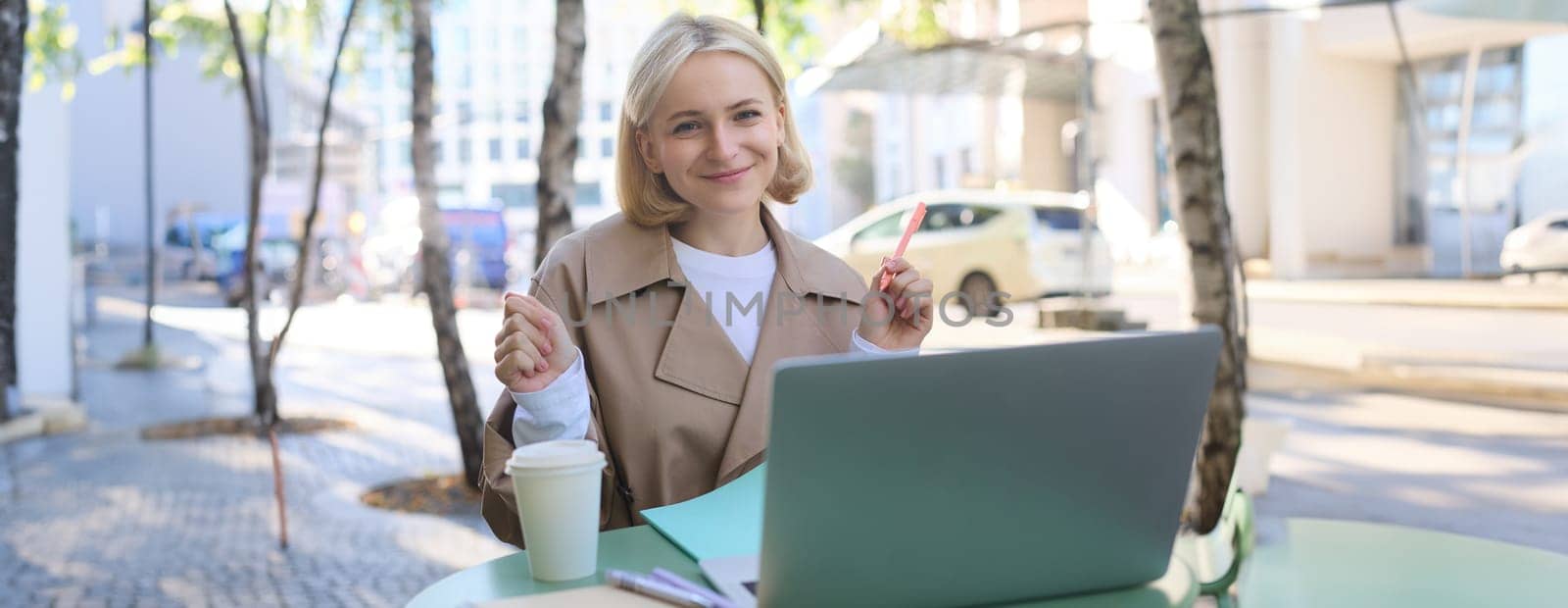 Remote workplace. Smiling young woman freelancer, student using laptop in outdoor cafe, talking to someone, attends online lecture, drinking coffee.