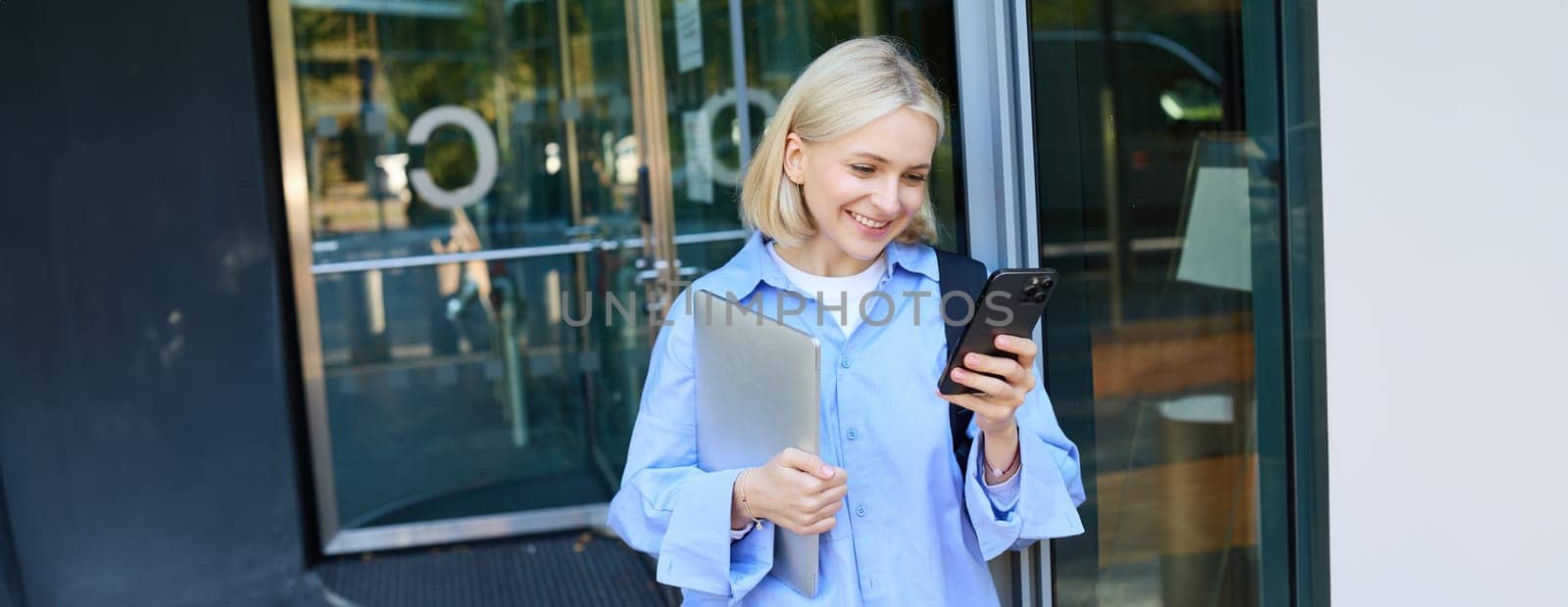 Image of young blond woman in blue shirt, holding laptop, waiting for someone near office building, using smartphone, mobile phone application.