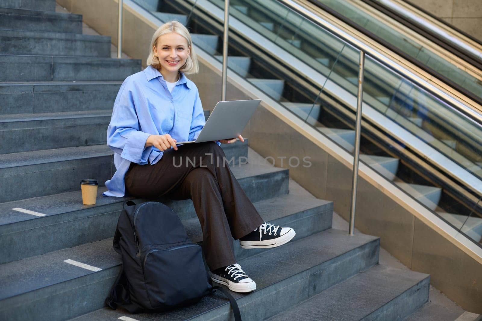 Image of stylish young modern woman, student doing homework, studying outdoors on campus stairs, sitting with laptop and coffee, drinking her cappuccino and connecting to public wifi by Benzoix