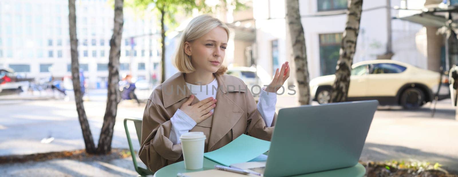Image of woman talking, introducing herself during online meeting, using laptop in outdoor cafe, working remotely, drinking coffee.